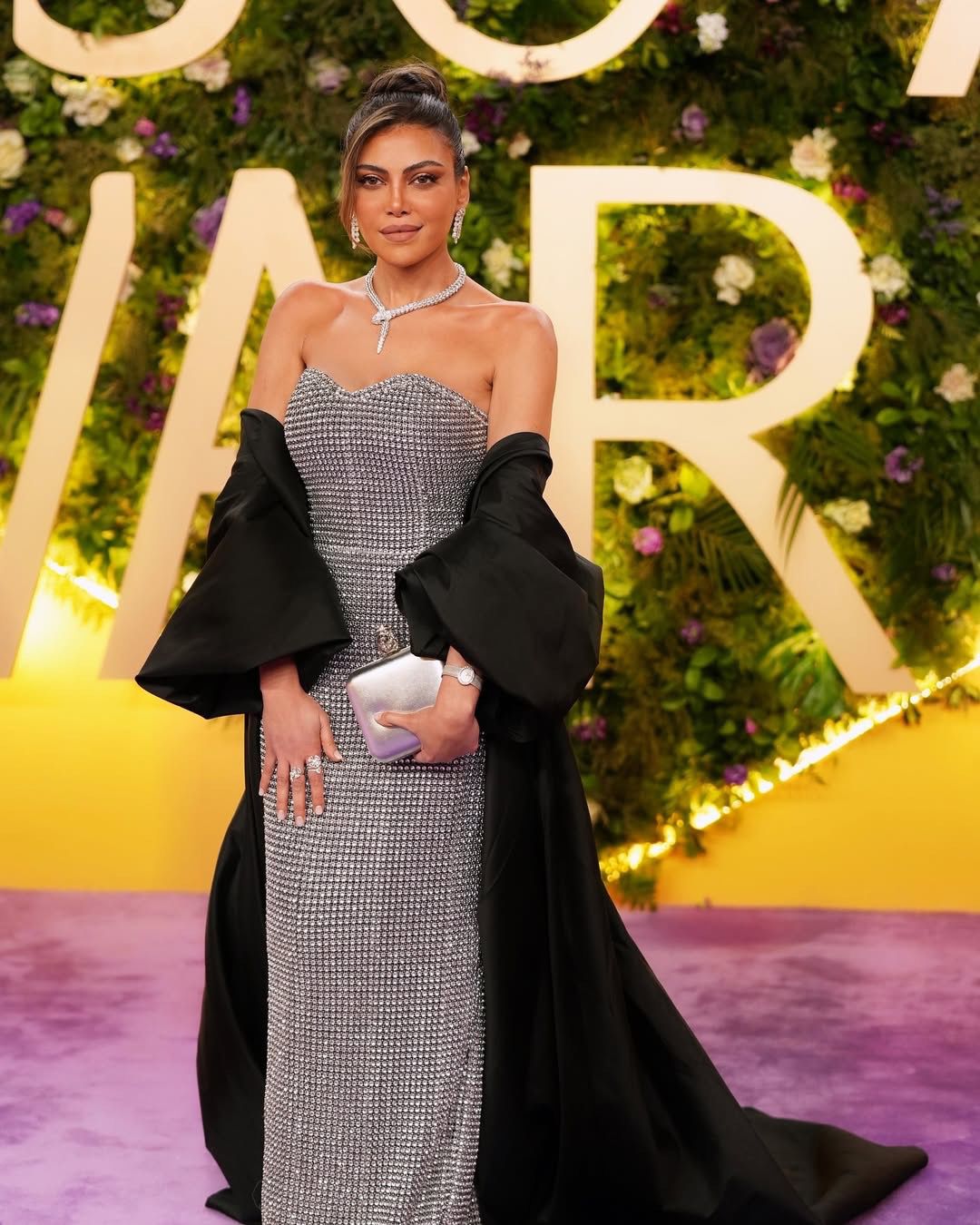 Woman in an elegant silver strapless dress with diamonds, black cape, and clutch on red carpet